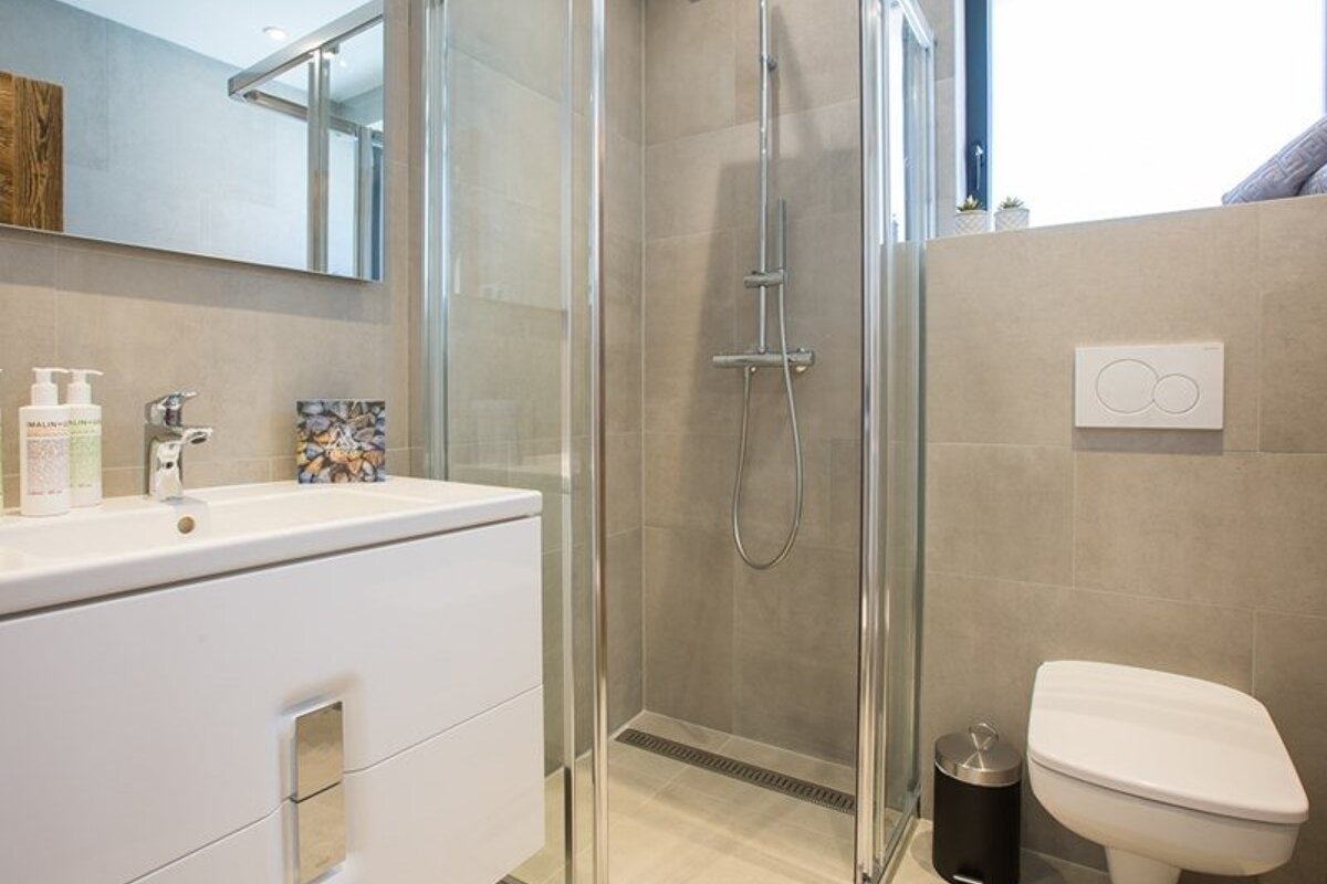 A modern bathroom with beige tiles, featuring a white vanity sink, a glass-enclosed shower, and a toilet.