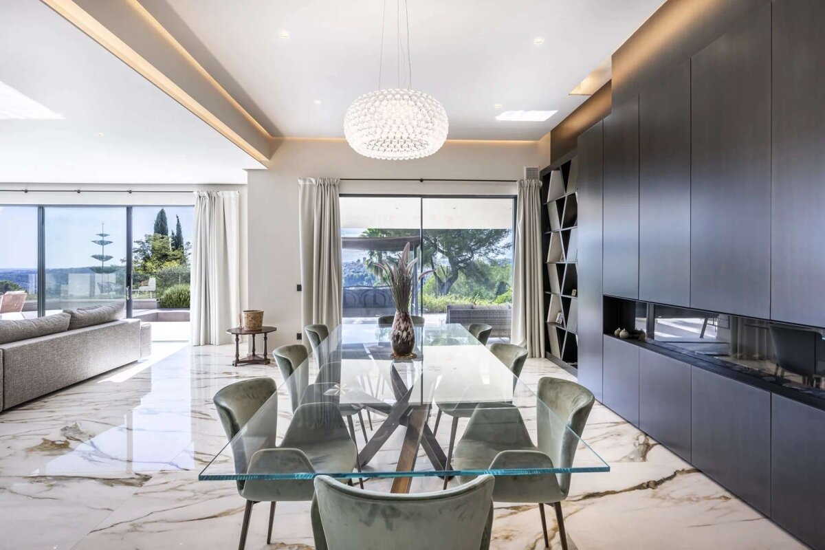 A luxurious, modern dining room with a glass table, plush chairs, sparkling chandelier, marble floor, and panoramic views through large windows.