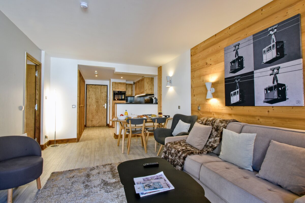 Modern alpine living room featuring a grey sofa, wood-paneled wall with cable car artwork, and a dining area in the background.