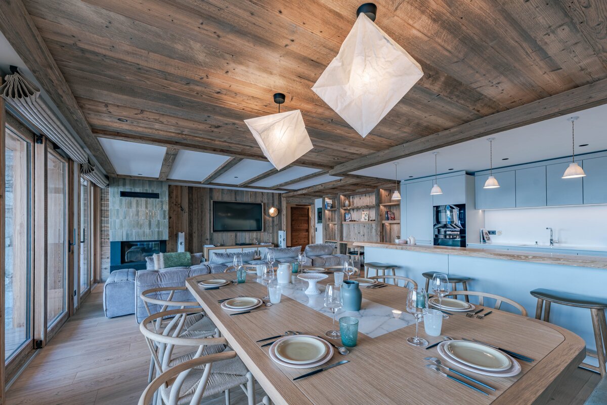 Open-plan chalet living space with a long dining table, light blue kitchen, rustic wood walls/ceiling, fireplace, and large windows.
