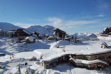 Panoramic view of a sunlit, snow-covered mountain ski resort with rustic wooden buildings nestled in vast peaks under a brilliant blue sky.