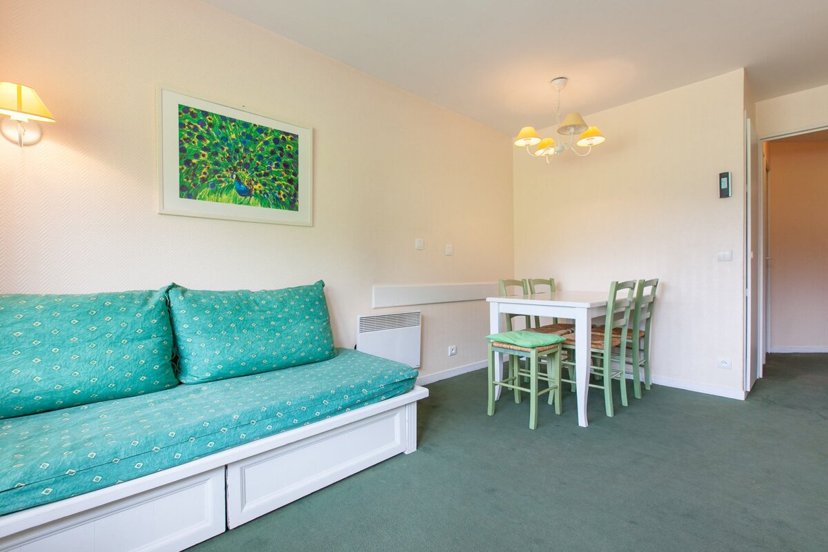 A living room with a turquoise sofa bed, green carpet, white dining table with light green chairs, and peacock art on the wall.