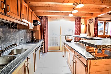 A rustic wooden kitchen with granite countertops, red curtains, and a bright window overlooking snowy chalet roofs. Ski decor suggests an alpine setting.