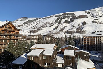 A snowy mountain ski resort featuring wooden chalets and apartment buildings, with visible ski slopes on the mountain under a clear blue sky.