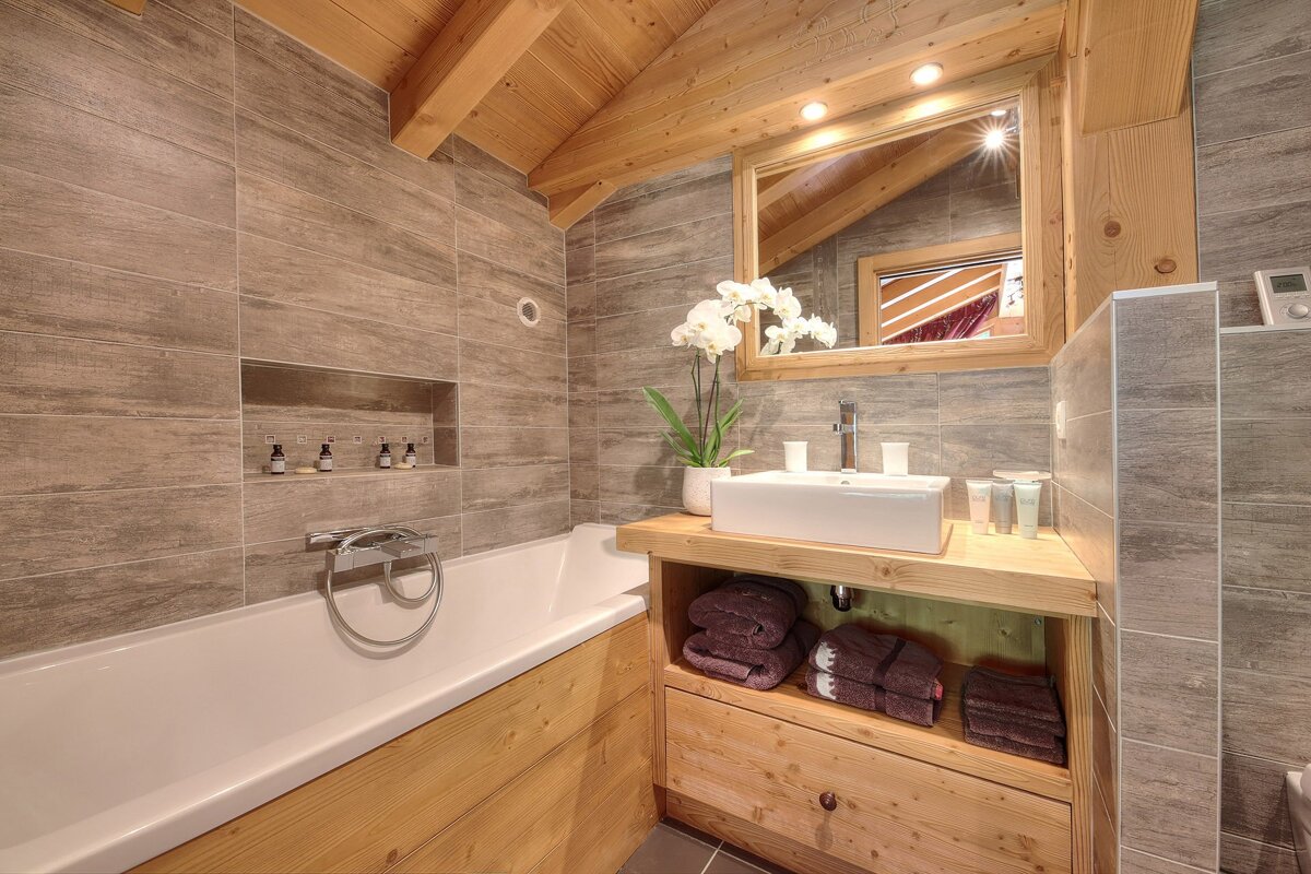 A modern, rustic bathroom featuring warm wood accents on the sloped ceiling, vanity, and tub surround, complemented by gray wood-look tiled walls. It includes a white tub, vessel sink, mirror, and an 