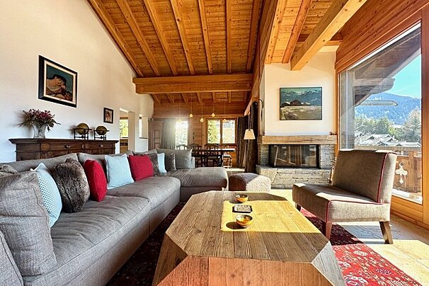 Cozy chalet living room with exposed wood ceilings, a large sectional sofa, wooden coffee table, fireplace, and scenic mountain views.
