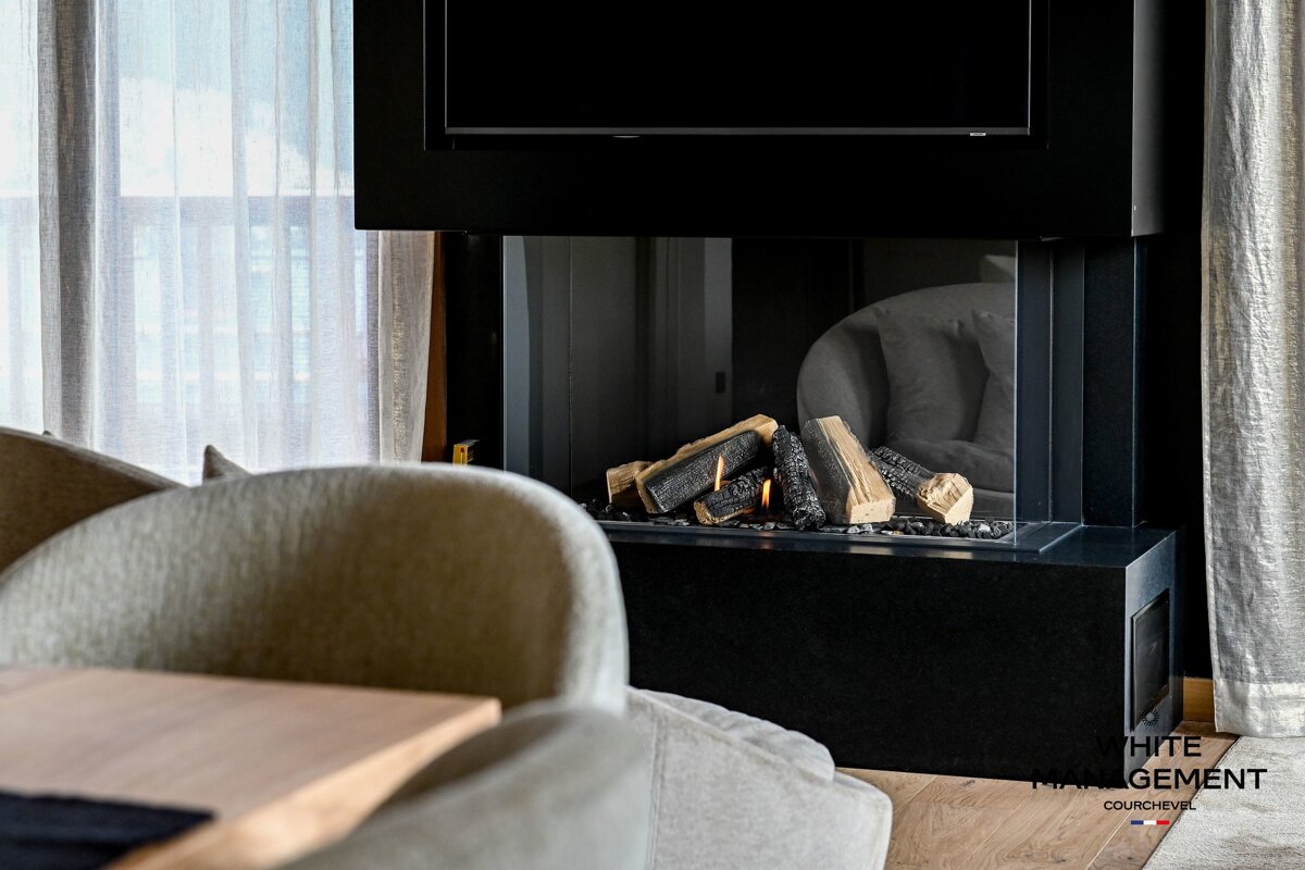 A modern living room features a black, glass fireplace with realistic burning logs and a TV above. Soft chairs and sheer curtains create a cozy atmosphere.