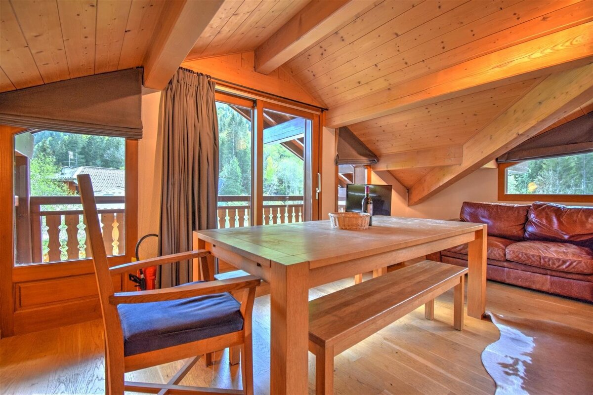 A warm, wood-paneled alpine room with a dining table, leather couch, and balcony overlooking lush trees. A high, beamed ceiling completes the cozy feel.