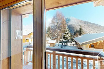 A view of a snow-covered mountain village with pine trees, as seen through a wooden balcony doorway.