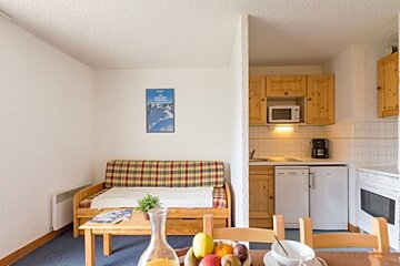 A cozy studio apartment with a plaid sofa bed, coffee table with fruit, and an open kitchenette. A ski poster hangs on the wall.