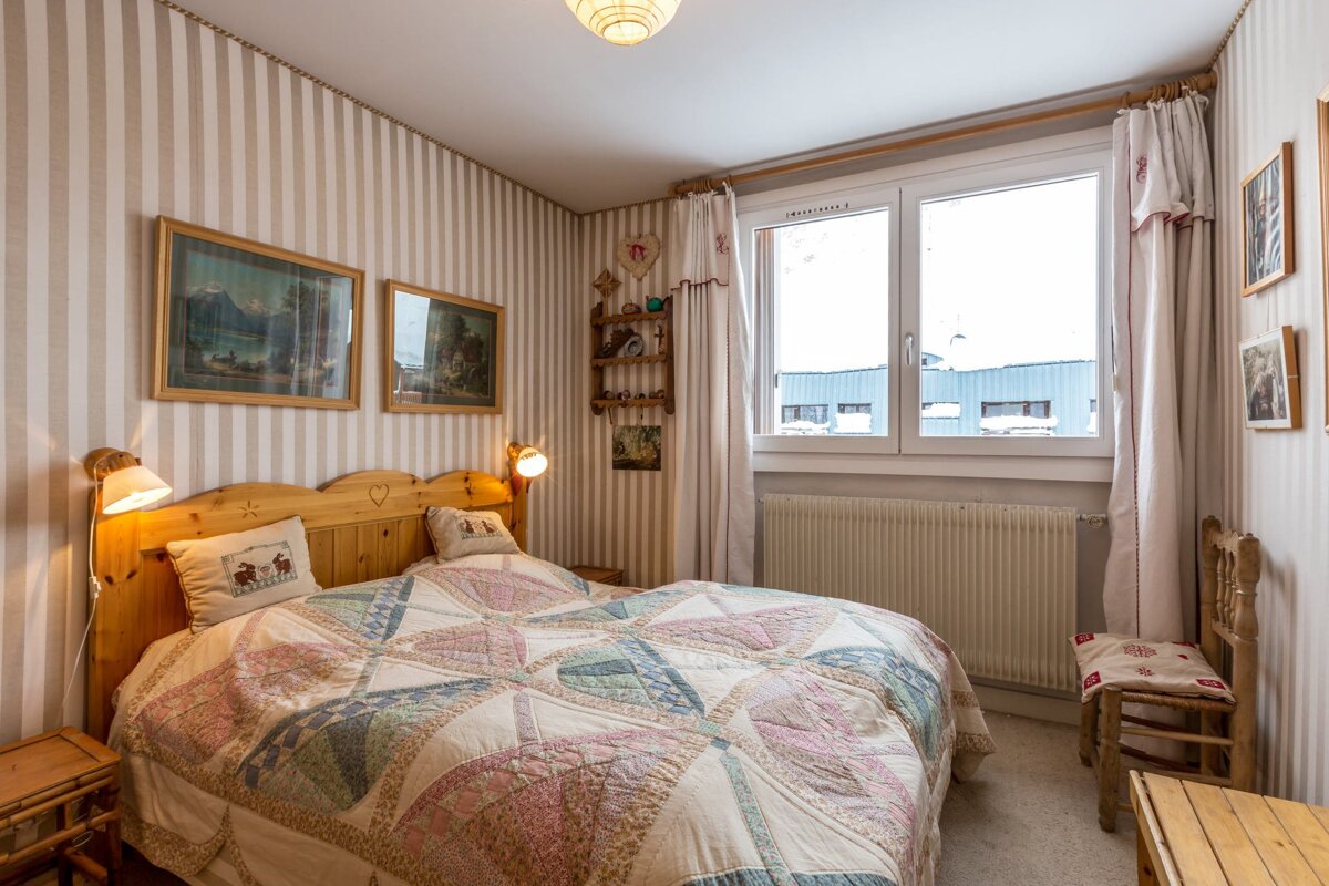 Cozy bedroom with a wooden bed, patterned quilt, striped wallpaper, framed pictures, and a large window looking out to snowy rooftops.