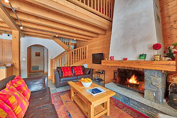 Cozy chalet living room with warm wood paneling, exposed beams, a crackling stone fireplace, leather sofas, and a wooden coffee table.