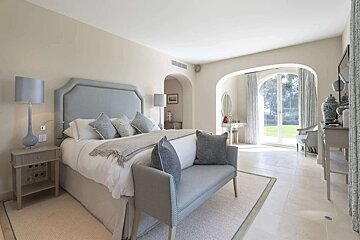 A luxurious, serene bedroom with a large bed, blue headboard, and matching bench. Arched glass doors open to a lush green garden.