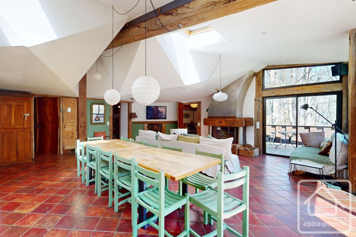 Spacious, rustic-modern open-plan room with a long dining table, red tiled floor, exposed beams, fireplace, and large windows looking out to trees.