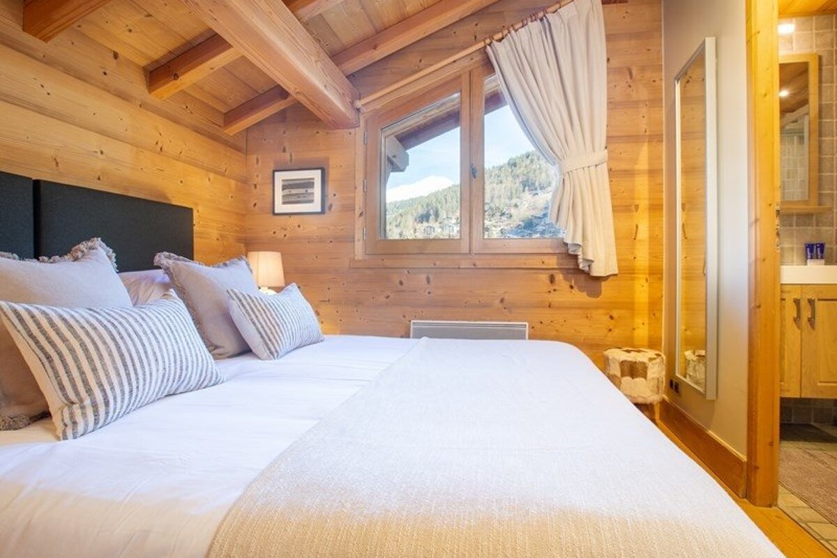 Cozy chalet bedroom with wooden walls, a large bed with striped pillows, and a window showing mountain views.
