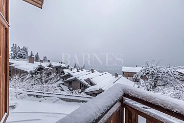 A snow-covered village of chalets and trees under an overcast sky, viewed from a snowy balcony railing, depicting a winter wonderland.
