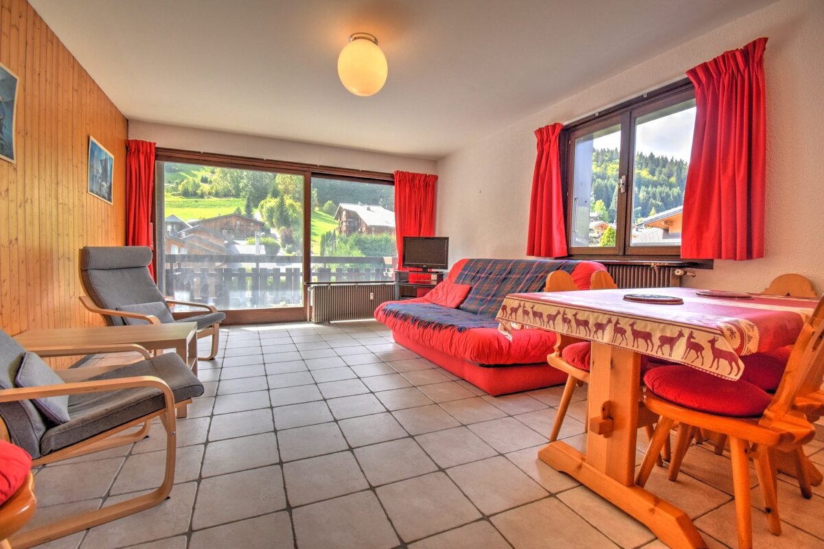 Cozy living/dining room with a red sofa, rustic wooden table, armchairs, and tiled floor. Large windows offer a view of a green, mountainous village.