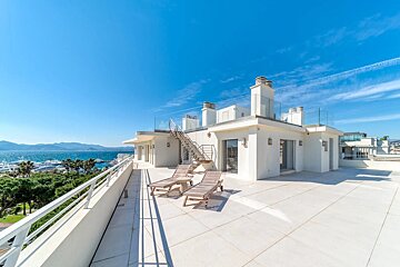 Modern white rooftop terrace with sun loungers overlooking a vibrant marina, blue sea, and distant mountains under a clear blue sky.