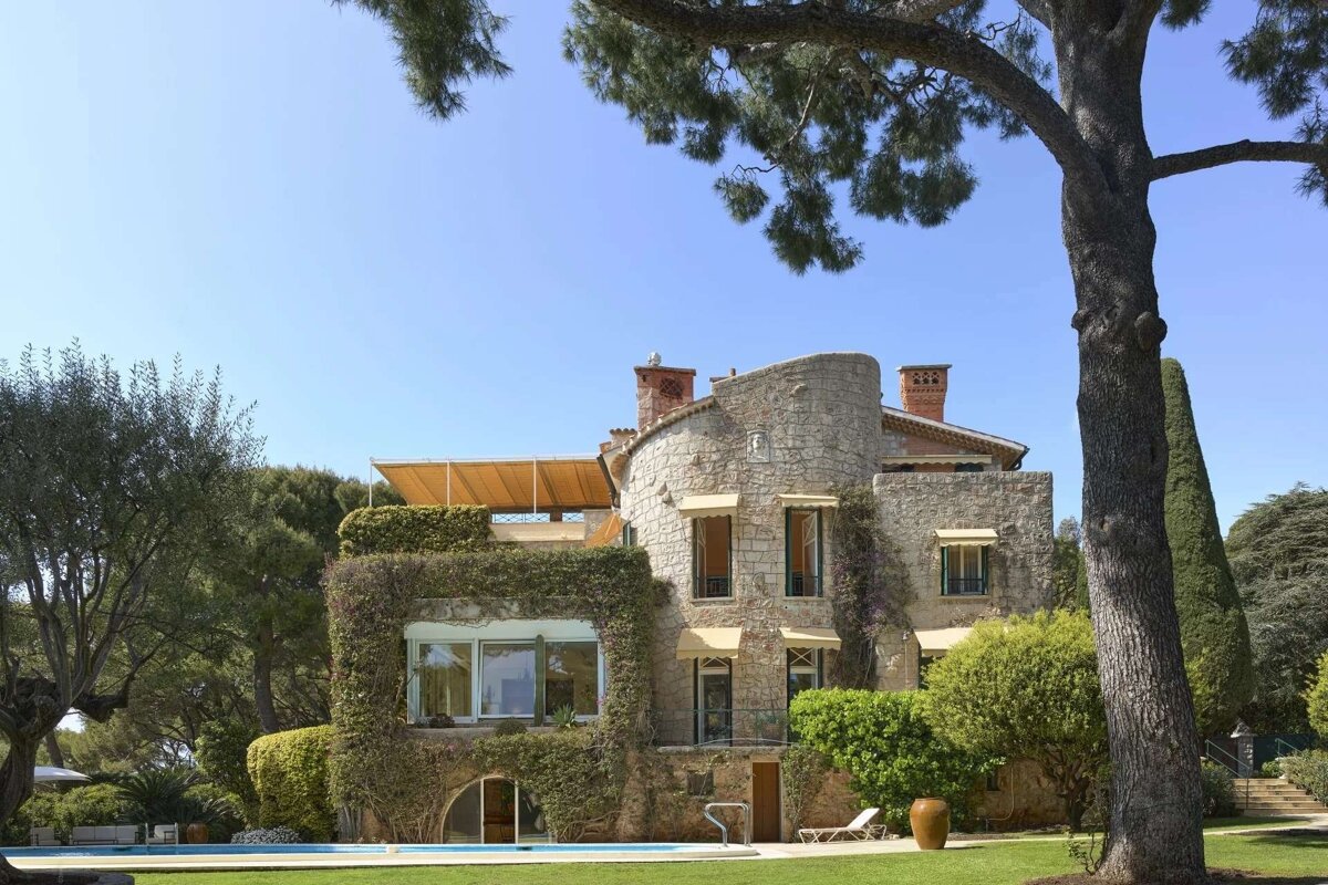 A grand stone villa covered in ivy, with a swimming pool and manicured gardens under a clear blue sky, framed by a large tree.