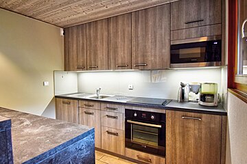 A modern kitchen featuring wood-grain cabinets, dark countertops, built-in appliances (oven, microwave), under-cabinet lighting, and a wooden ceiling.