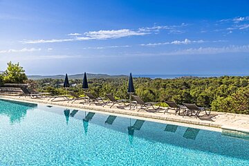 An infinity pool with loungers and umbrellas overlooks a vast green landscape and the distant sea under a bright blue sky.
