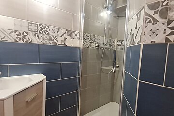 A modern bathroom features a shower cabin, sink, and walls tiled in blue, grey, and a decorative band of intricate patterned tiles.