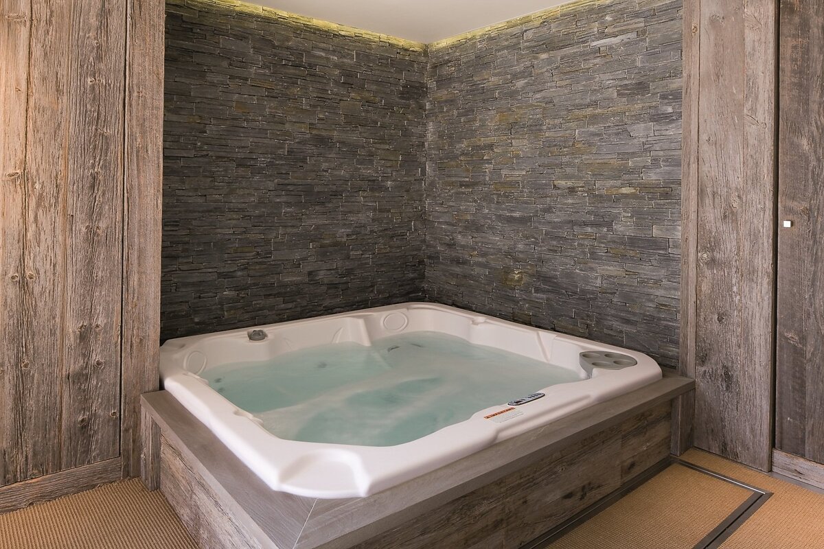 A white hot tub with bubbling water is set in a cozy room featuring textured dark stone walls, rustic wooden panels, and subtle overhead lighting.