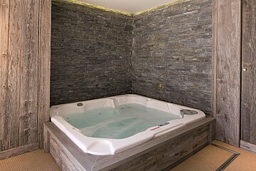 A bubbling white hot tub is surrounded by dark slate stone walls and weathered wooden panels, lit by warm ceiling lights.