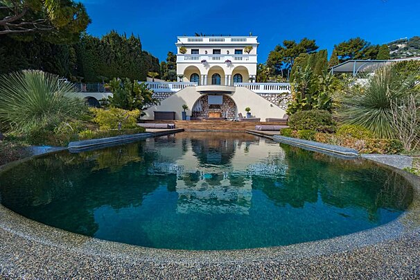 A grand villa with arched balconies overlooks a large, dark-bottomed swimming pool, reflecting the house and surrounded by lush green landscaping under a clear blue sky.