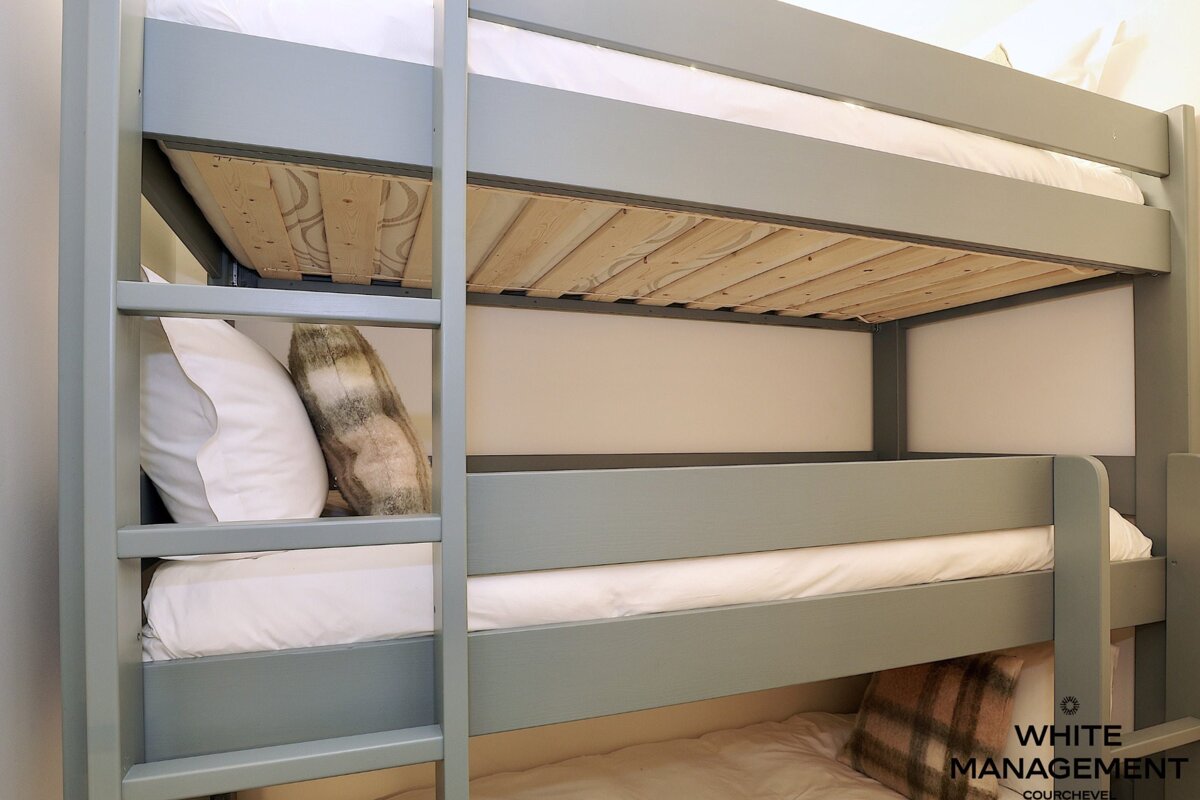 A grey wooden bunk bed with multiple tiers, white bedding, plaid pillows, and an integrated ladder.
