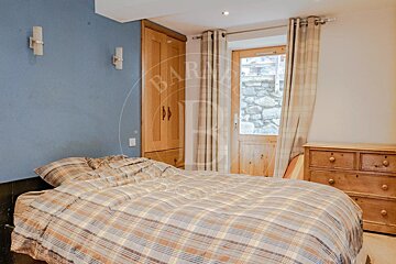 A cozy bedroom featuring a plaid bed, blue wall, wooden wardrobe, dresser, and a rustic door with a stone view outside.