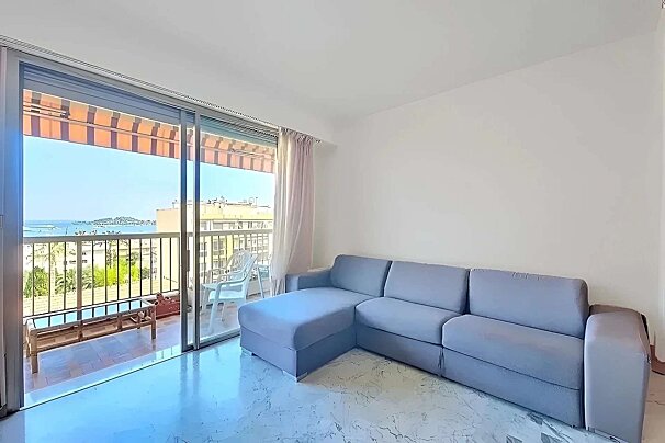 A clean living room with a grey sectional couch and white walls, featuring a balcony with an orange awning overlooking the sea.