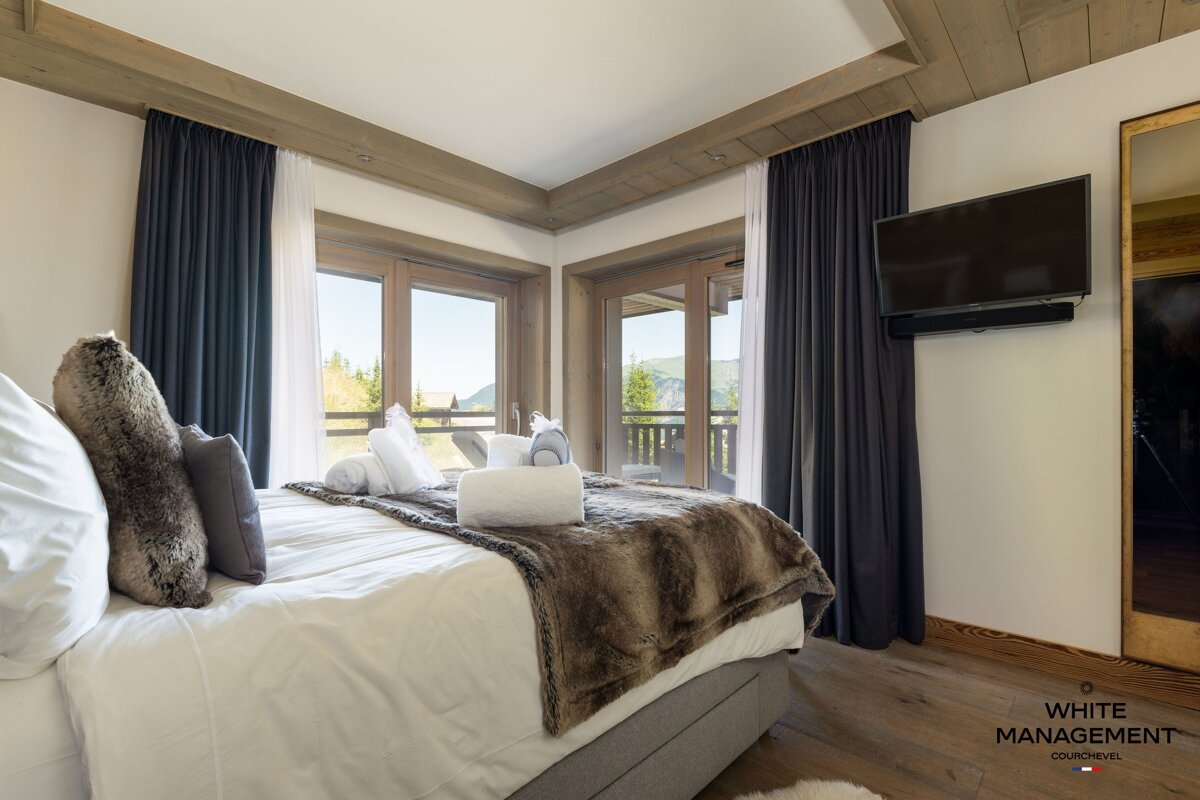 Cozy chalet bedroom featuring a bed with a fur throw, wood ceilings, large windows with scenic mountain views, and a wall-mounted TV.