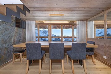 Rustic modern dining room with a wooden table, grey chairs, wood paneling, and a stone wall, offering a panoramic view of snow-capped mountains.