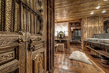 An ornate carved door opens to a rustic wooden bathroom with a white sheepskin rug and double vanity sinks.