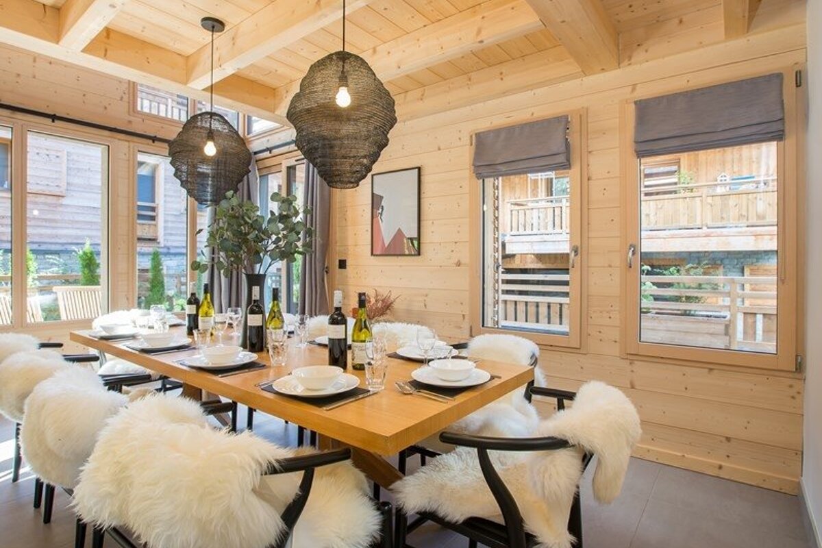 A chalet dining room featuring a wooden table set for dinner, chairs with white sheepskin throws, and large dark pendant lights.