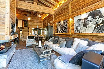 A luxurious, rustic chalet interior with rich wooden walls and ceiling, a large comfy sofa, a modern fireplace, and oversized black and white artwork.