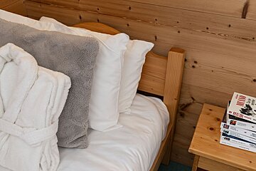 A cozy wooden bed with white and grey pillows, a folded white bathrobe, and a stack of books on a wooden nightstand.