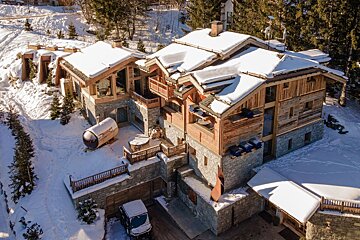 Luxurious, multi-level wooden and stone chalet covered in fresh snow on a sunny winter day, with trees, barrel sauna, and hot tubs visible.