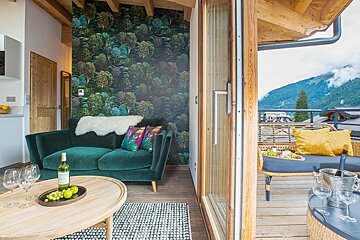 Cozy chalet living room with a green velvet sofa and forest wallpaper next to a wooden balcony with mountain views.