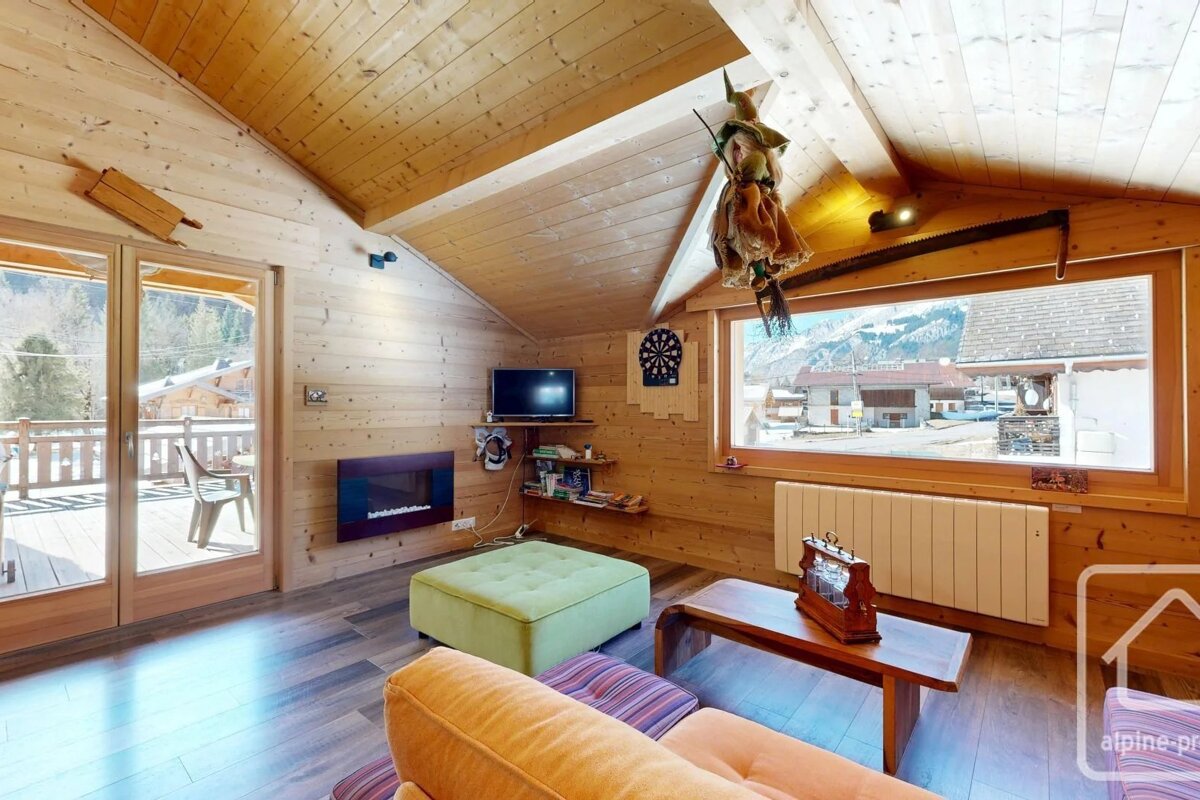 Cozy wooden chalet living room featuring a green ottoman, a fireplace, and scenic window views of a mountain village.