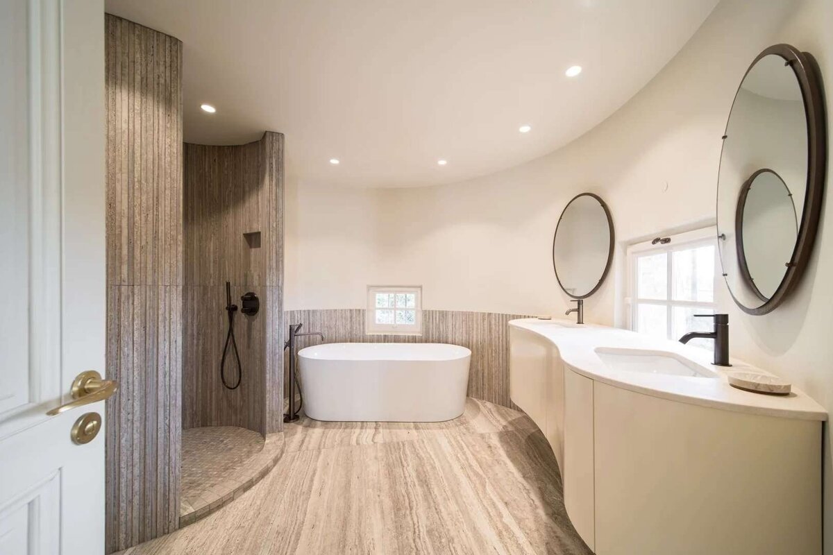 A contemporary bathroom with elegant curved architecture, featuring a walk-in shower, freestanding tub, and a double vanity with round mirrors in natural tones.