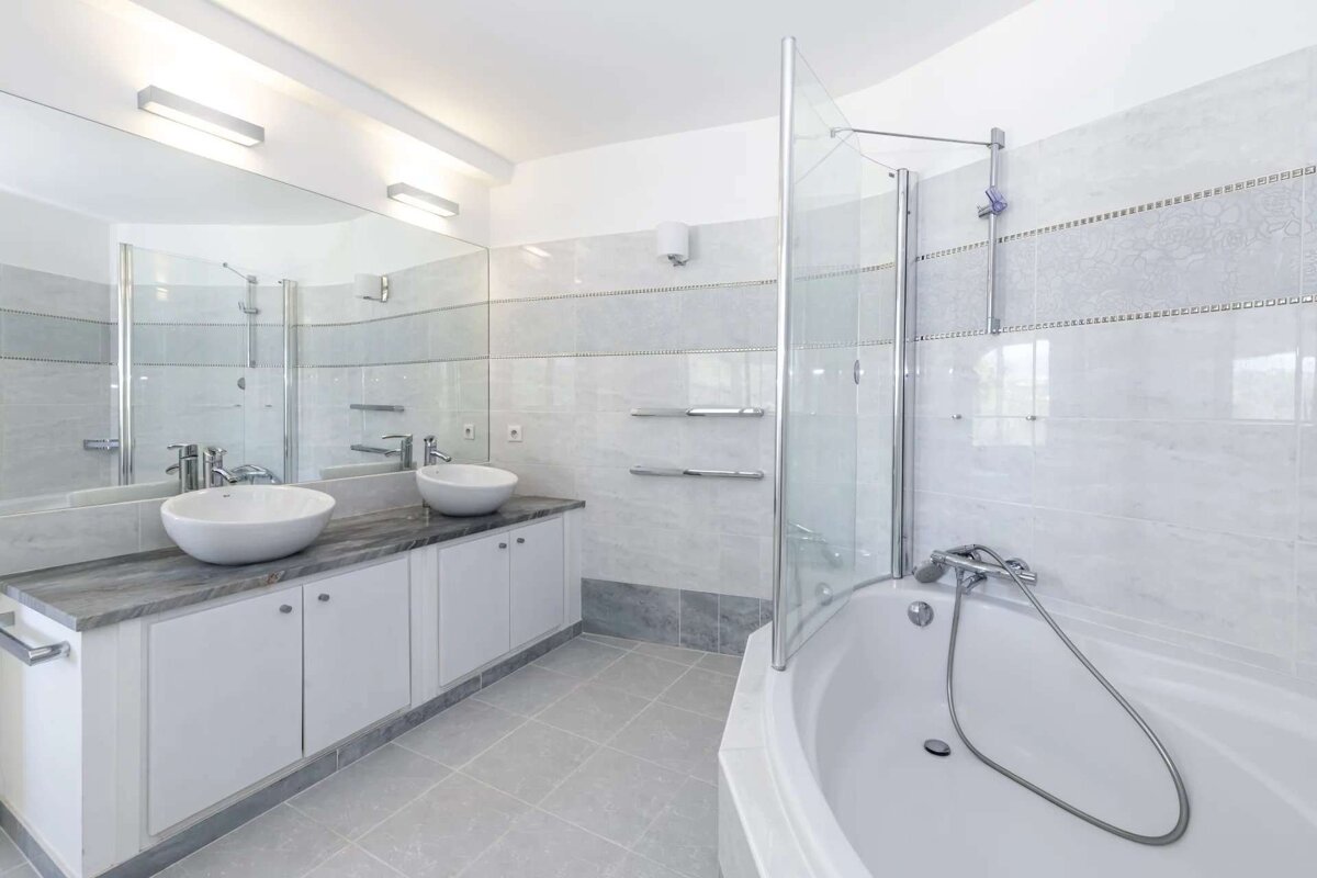 A clean, modern bathroom with a double vanity, vessel sinks, a large mirror, and a corner bathtub with a glass shower. Grey and white tiles.