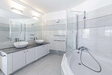 A clean, modern bathroom with a double vanity, vessel sinks, a large mirror, and a corner bathtub with a glass shower. Grey and white tiles.