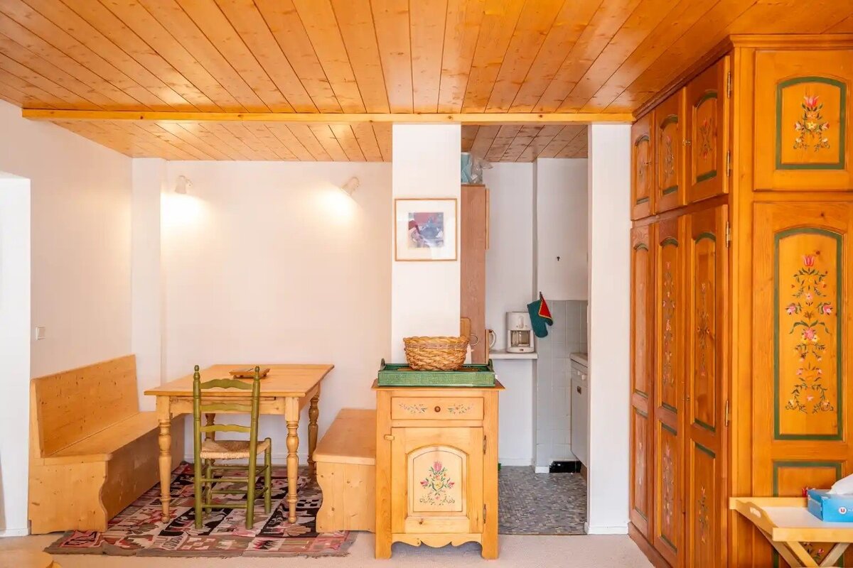 A rustic room with a wooden ceiling, dining table & benches, and a kitchenette. Features traditional, painted wooden cabinets.