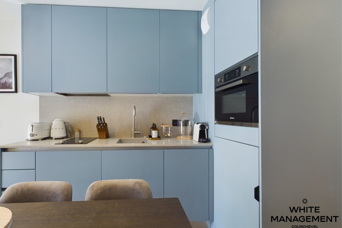 A modern kitchen featuring light blue cabinets, beige countertops, a built-in oven, and a dining table in the foreground.