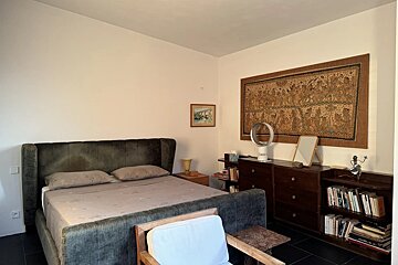 A cozy bedroom with a dark velvet upholstered bed, wooden dressers, a bookcase, wall art, and a chair in the foreground.