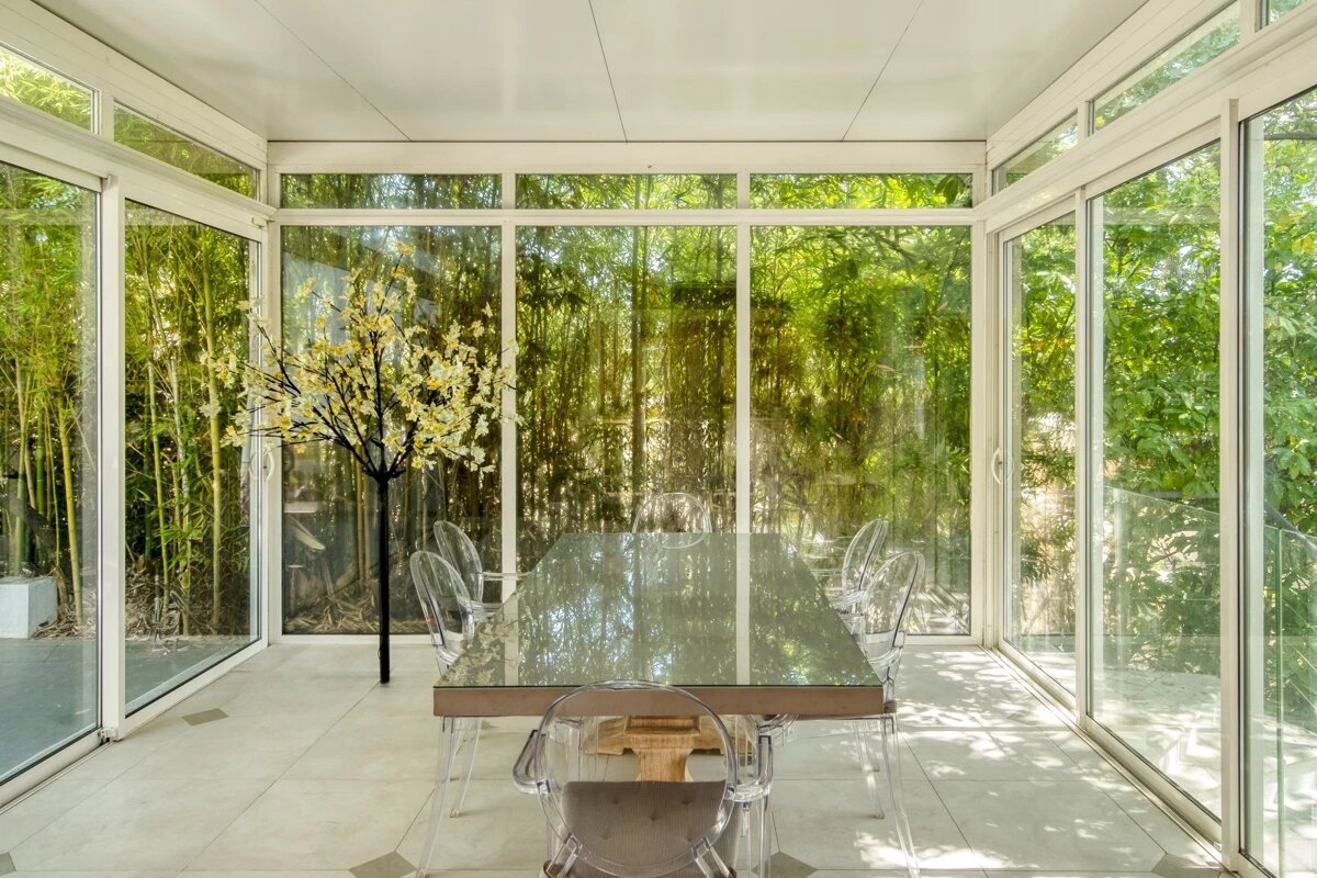 Bright sunroom with a glass-topped dining table, clear acrylic chairs, and floor-to-ceiling windows overlooking lush green bamboo.