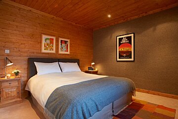 A cozy bedroom with warm wooden walls/ceiling, a bed with white linens and blue throw, two nightstands with lamps, framed art, and a colorful rug.