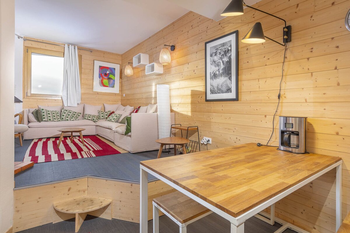 A cozy room with light wood paneling, featuring a raised living area with a large sofa and a red rug, and a lower dining space with a wooden table.